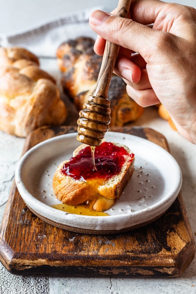 slice of challah bread with jam spread on top and a drizzle of honey, a hand holding a spoon of honey on top of the toast.