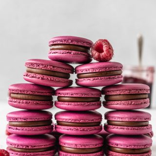 pink macarons with chocolate filling stacked with raspberries on top.