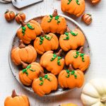 Pumpkin Macarons shaped like pumpkins, on a plate seen from the top with pumpkins around and a sign saying harvest.
