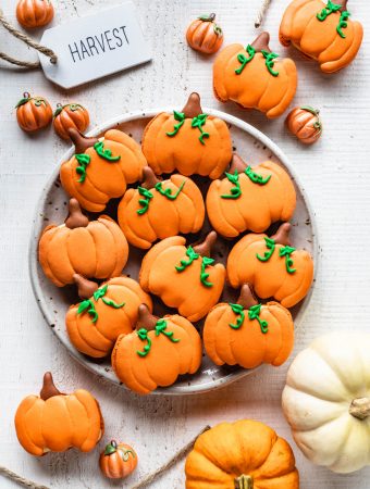 Pumpkin Macarons shaped like pumpkins, on a plate seen from the top with pumpkins around and a sign saying harvest.
