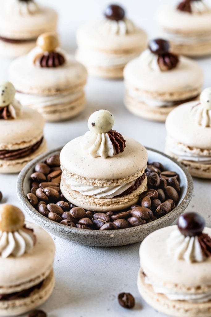 coffee macaron on top of a bowl full of coffee beans, more coffee macarons around it, all macarons have a chocolate covered espresso bean on top.