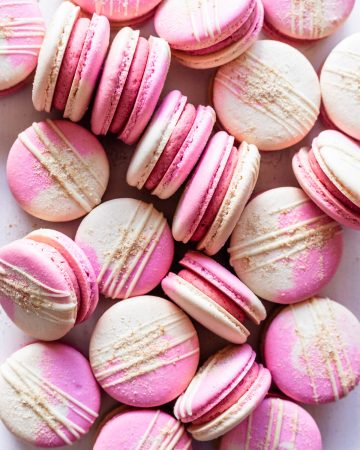 Strawberry macarons with two color shells drizzled with white chocolate and topped with graham cracker crumb.