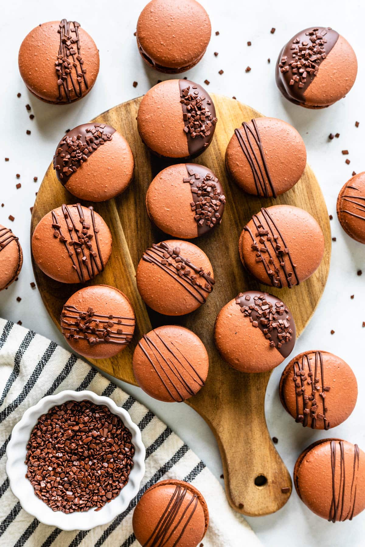 chocolate macarons filled with chocolate buttercream on a wooden board, drizzled with chocolate, or dipped in chocolate and topped with sprinkles, seen from the top