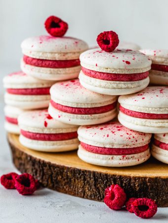 vegan macarons filled with pink raspberry frosting topped with speckles, surrounded by freeze dried raspberries.