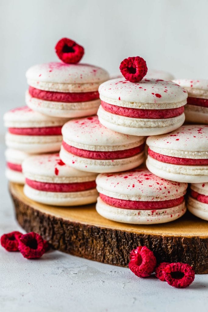 vegan macarons filled with pink raspberry frosting topped with speckles, surrounded by freeze dried raspberries.