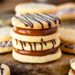 Finished alfajor cookies filled with dulce de leche and drizzled with chocolate, arranged on a wooden board.
