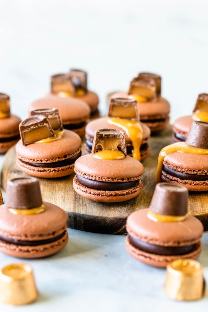 Chocolate Caramel Macaron in the front filled with caramel and chocolate topped with a rolo candy on a wooden board, with more macarons around it