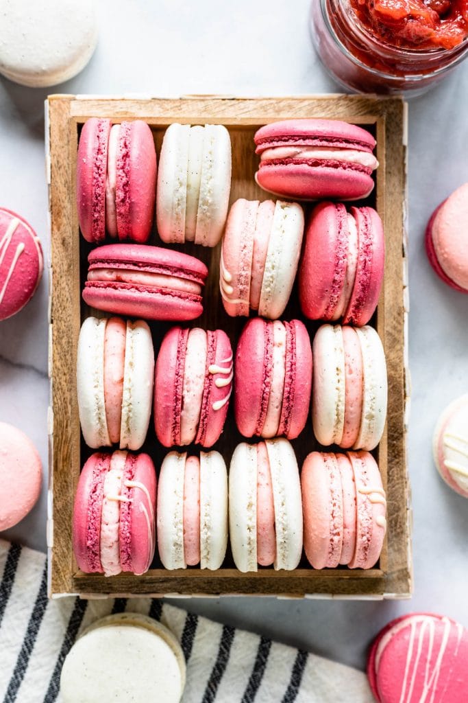 Pink, white, and dark pink strawberry rhubarb macarons in a box, from a bird's eye view, with jam in the top right corner
