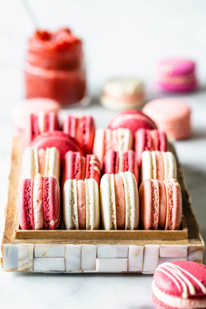 Pink, white, and dark pink strawberry rhubarb macarons in a box, with jam in the top left corner