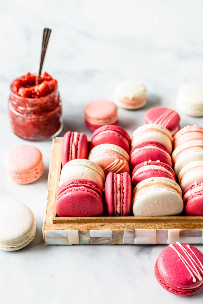 Pink, white, and dark pink strawberry rhubarb macarons in a box, with jam in the top left corner