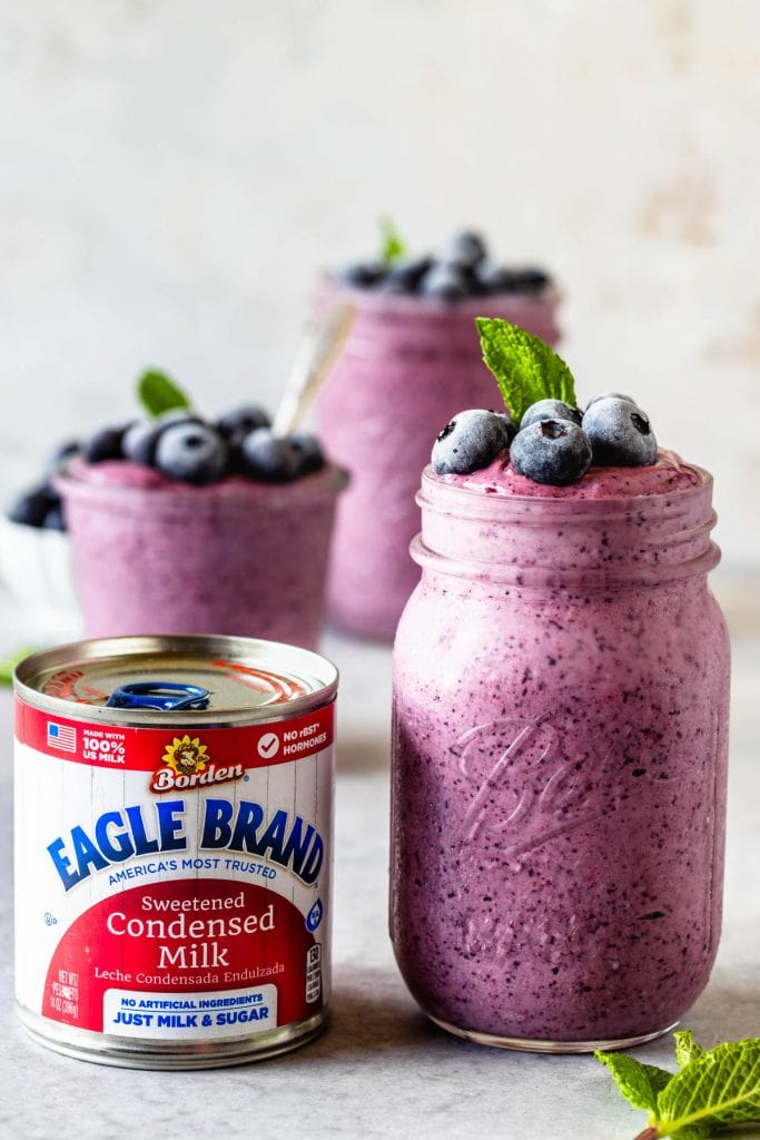 can of condensed milk and blueberry slushie in a mason jar topped with mint leaf and frozen blueberries and a spoon