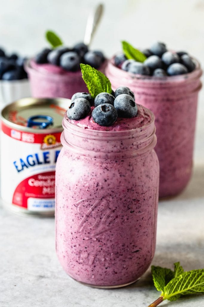 can of condensed milk and blueberry slushie in a mason jar topped with mint leaf and frozen blueberries and a spoon