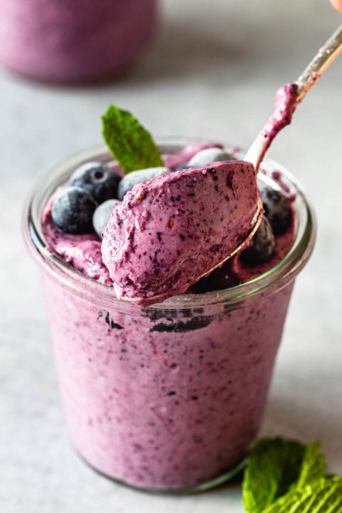 spoon scooping blueberry slushie in a mason jar topped with mint leaf and frozen blueberries and a spoon