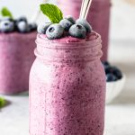 blueberry slushie in a mason jar topped with mint leaf and frozen blueberries and a spoon