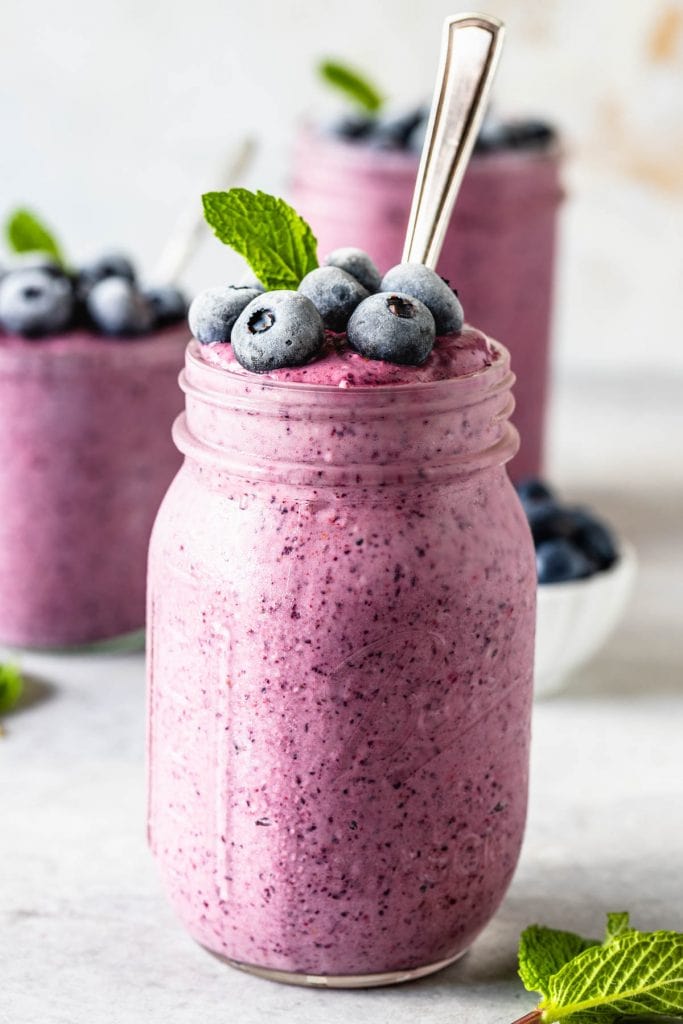 blueberry slushie in a mason jar topped with mint leaf and frozen blueberries and a spoon