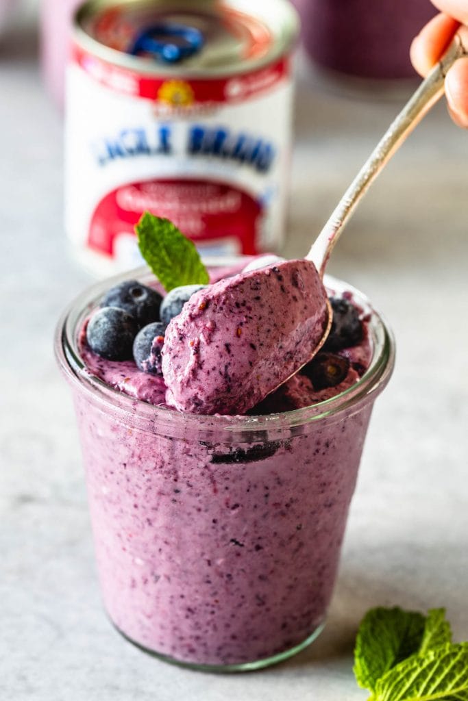 spoon scooping blueberry slushie in a mason jar topped with mint leaf and frozen blueberries and a spoon