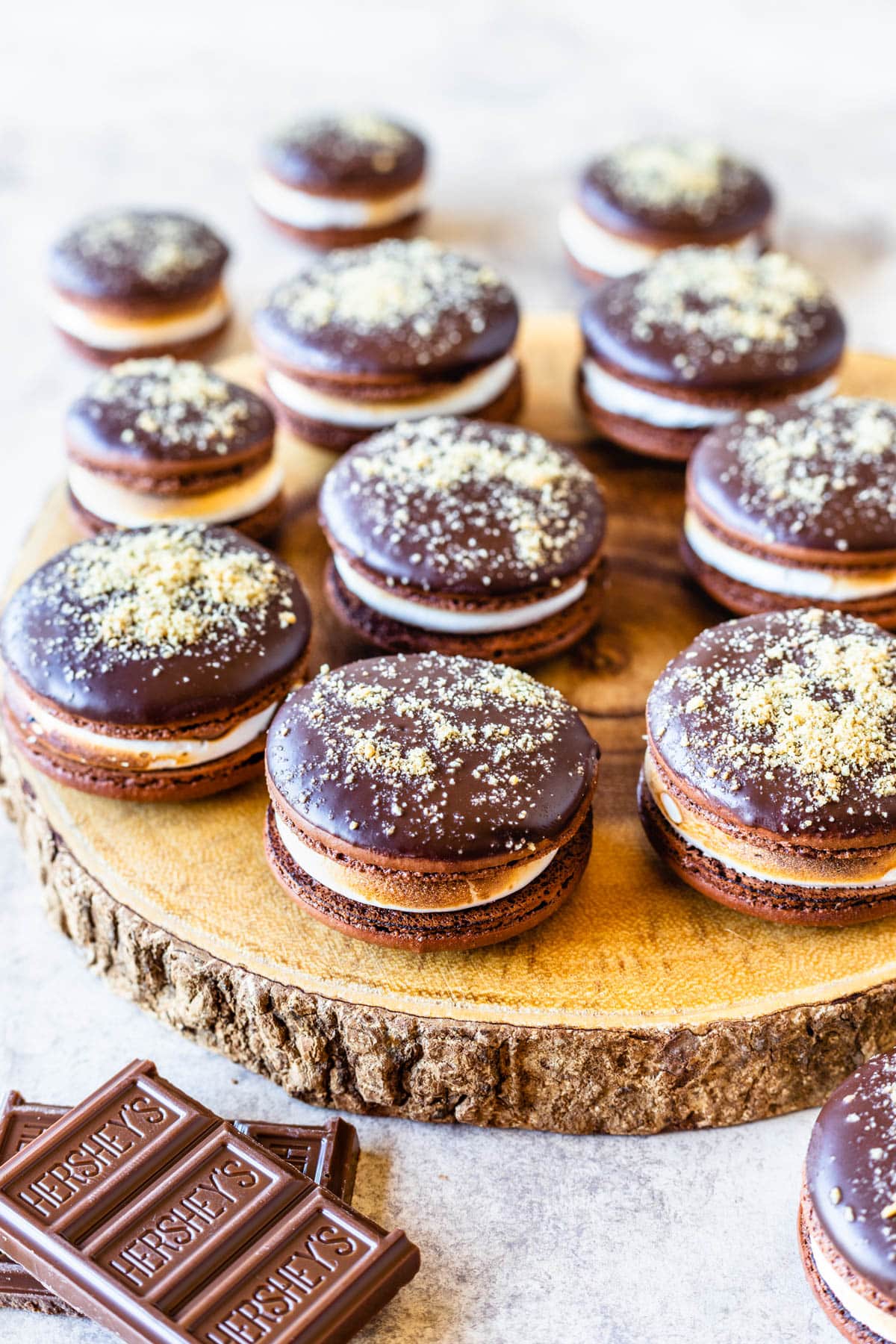 S'mores Macarons dipped in chocolate topped with graham cracker crumbs filled with toasted marshmallow on top of a wood board