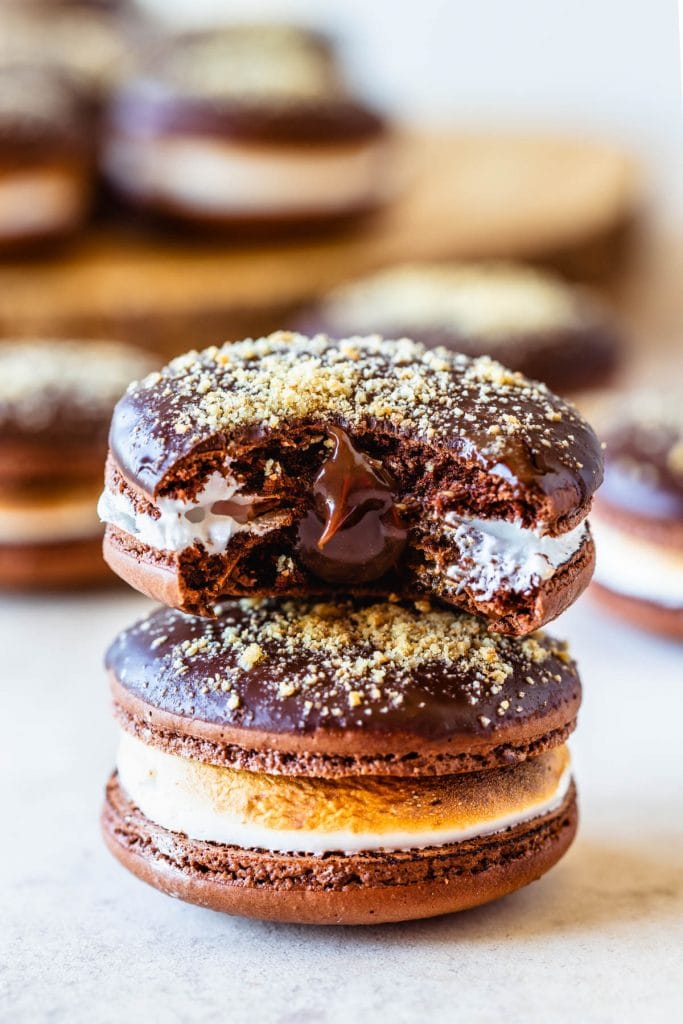 S'mores Macarons dipped in chocolate topped with graham cracker crumbs filled with toasted marshmallow stacked on top of each other with a bite taken out of the top cookie