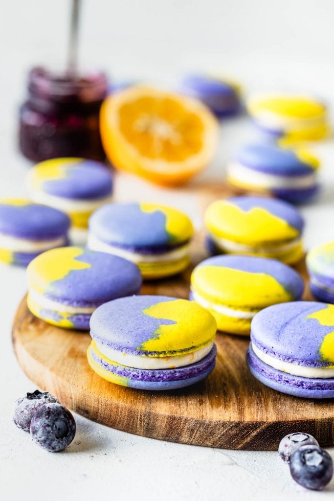purple and yellow vegan macarons filled with buttercream on top of a wooden board with blueberries around and on the back a jar of blueberry jam and half of a lemon