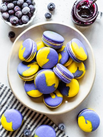 vegan macarons the shells are purple and yellow, they are filled with lemon buttercream and are in a white bowl with a container with blueberries and a container with jam around