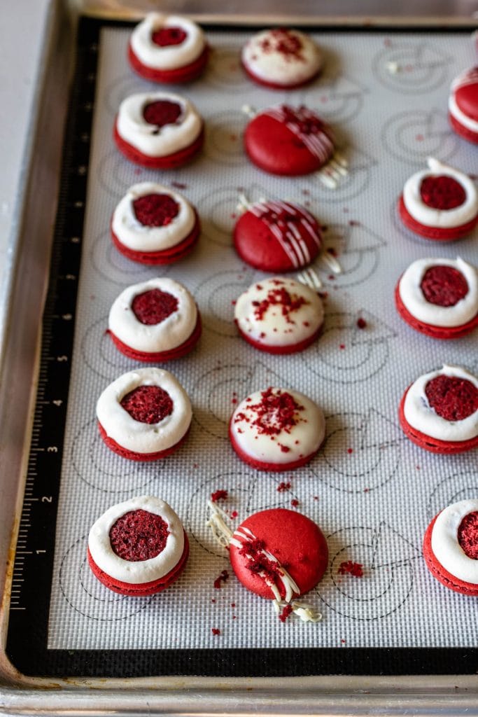 red macaron shells filled with red velvet cake and cream cheese frosting, and shells drizzled with white chocolate