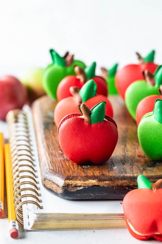macarons shaped like apples on top of a wooden board and a notebook with pencils on the side.