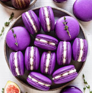 purple macarons filled with fig jam and buttercream seen from the top, with sprigs of thyme all around, and fresh figs on the top left corner and on the bottom left corner.