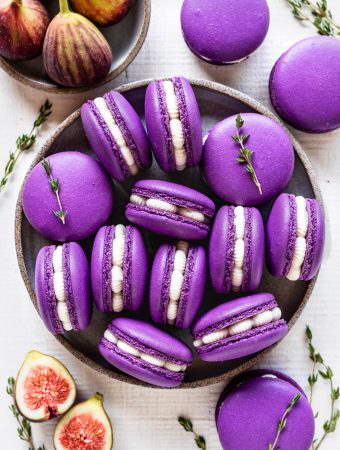 purple macarons filled with fig jam and buttercream seen from the top, with sprigs of thyme all around, and fresh figs on the top left corner and on the bottom left corner.
