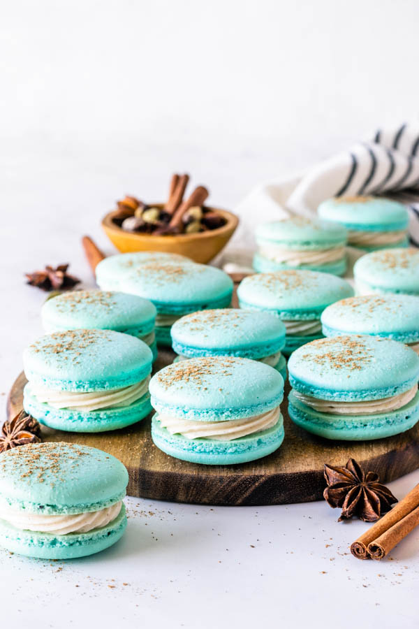 chai macarons filled with chai buttercream, on top of a wooden board, topped with sprinkled cinnamon, with cinnamon sticks and anise stars around.