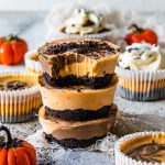 Chocolate Pumpkin Mini Cheesecakes stacked, the first cheesecake has a bite taken out of it. Around there are pumpkins and more mini cheesecakes decorating. they are topped with oreo crumbs and the cheesecakes on the back are topped with whipped cream.