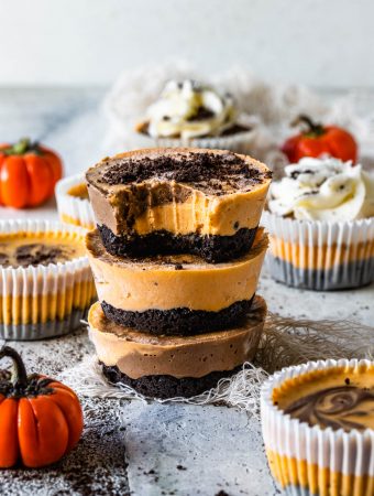 Chocolate Pumpkin Mini Cheesecakes stacked, the first cheesecake has a bite taken out of it. Around there are pumpkins and more mini cheesecakes decorating. they are topped with oreo crumbs and the cheesecakes on the back are topped with whipped cream.