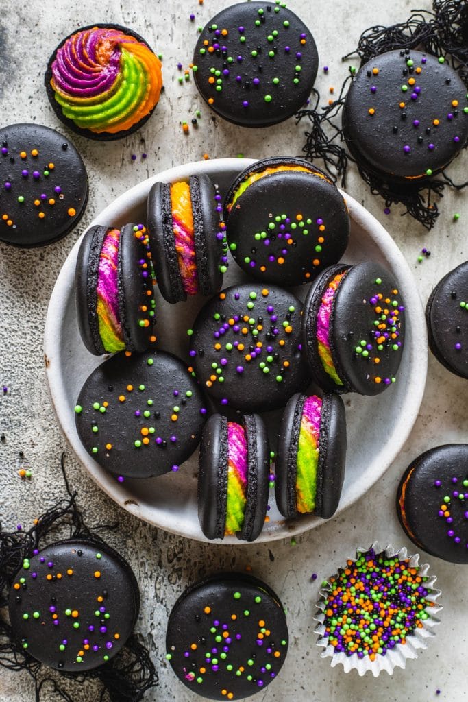 halloween Macarons filled with a purple, green and orange frosting, topped with colorful sprinkles, in a plate.