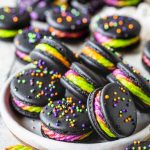 Black Macarons filled with a purple, green and orange frosting, topped with colorful sprinkles, in a plate.
