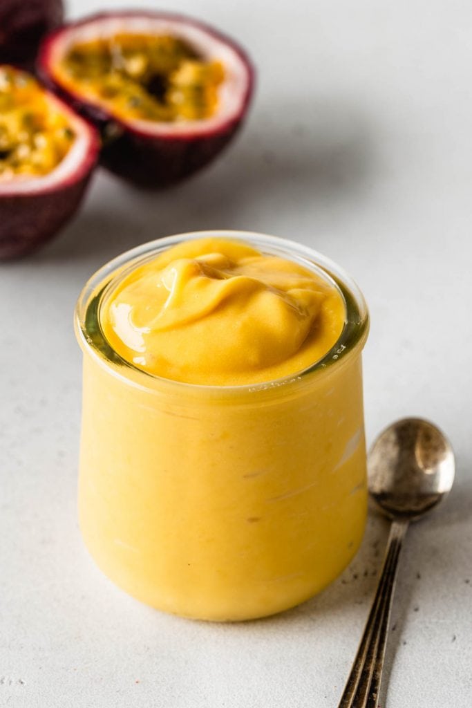 passion fruit curd in a glass container with a spoon on the side and fresh passionfruit on the back.