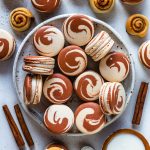 macarons with swirl shells filled with cinnamon frosting, on a plate, with a container of frosting on the side, with cinnamon rolls around.