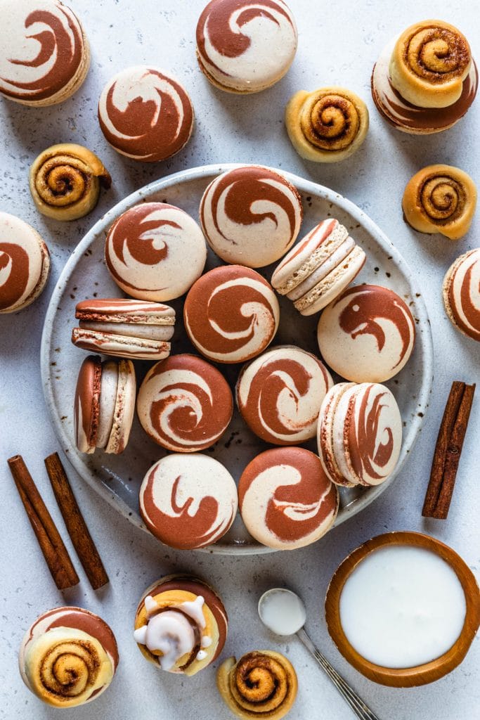 macarons with swirl shells filled with cinnamon frosting, on a place, with a container of frosting on the side, with cinnamon rolls around.