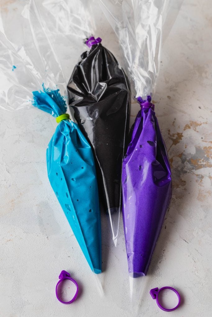three different color batters of macaron in three different piping bags: blue, black, and purple.