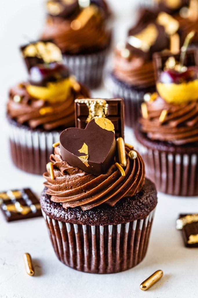 chocolate cupcakes filled with biscoff, topped with biscoff chocolate frosting, decorated with cherries dipped in gold, chocolate bars, chocolate hearts and gold sprinkles.