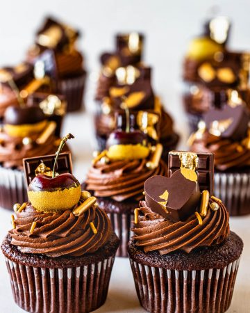chocolate cupcakes filled with biscoff, topped with biscoff chocolate frosting, decorated with cherries dipped in gold, chocolate bars, chocolate hearts and gold sprinkles.
