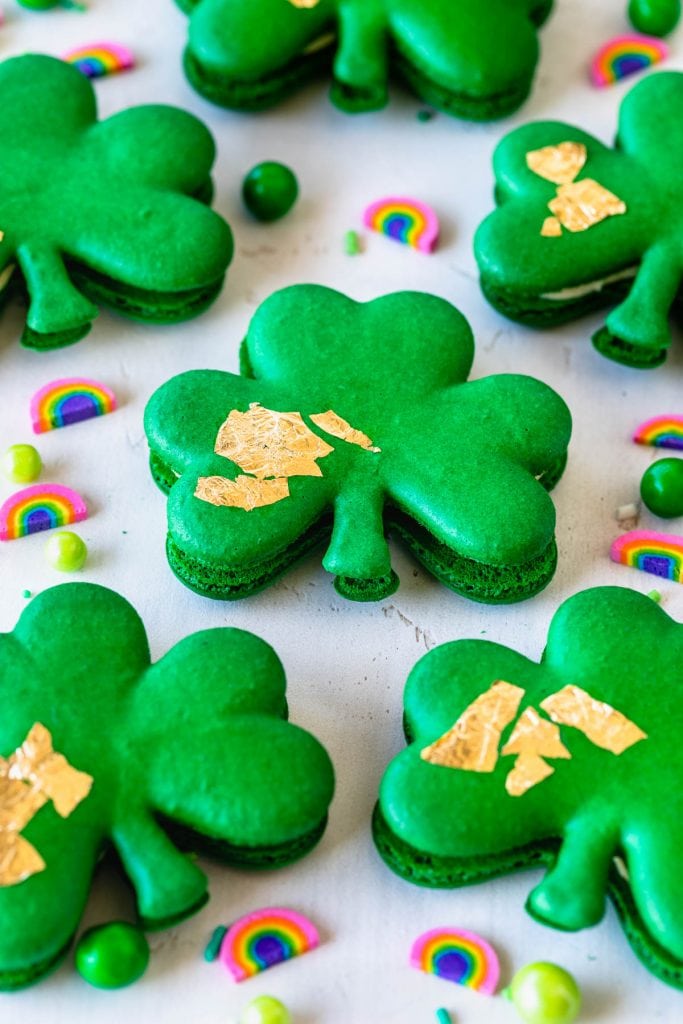 Shamrock macarons with gold edible leaf on top, surrounded by sprinkles.