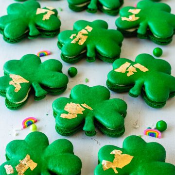 Shamrock macarons with gold edible leaf on top, surrounded by sprinkles.