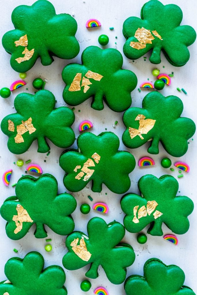 Shamrock macarons with gold edible leaf on top, surrounded by sprinkles.