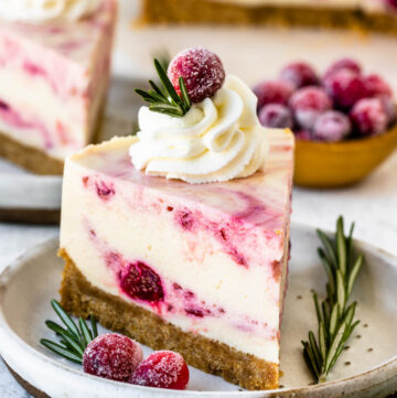 Cranberry cheesecake slice with whipped cream on top and sugared cranberries and a sprig of rosemary.