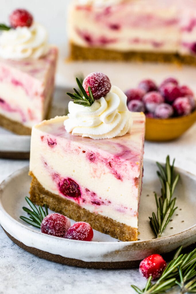 Cranberry cheesecake slice with whipped cream on top and sugared cranberries and a sprig of rosemary.