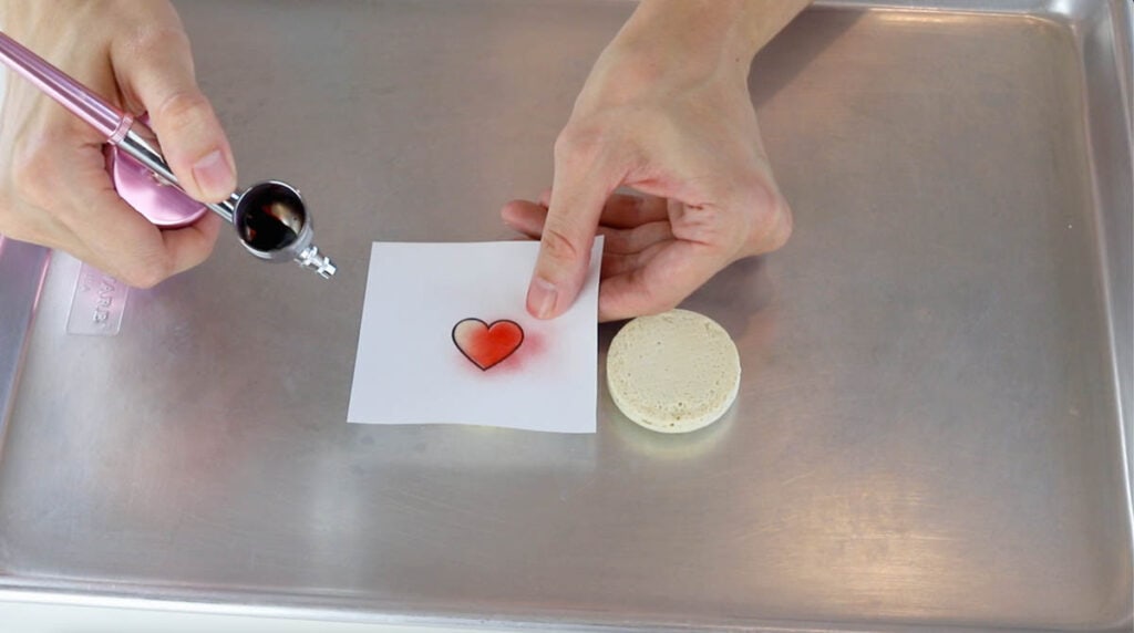 airbrushing the top of a macaron shell with a heart stencil.