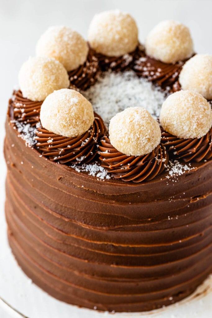 Coconut Chocolate Cake frosted with brigadeiro, topped with coconut fudge balls, with two mini cakes on the side, and brigadeiros spread around the table.