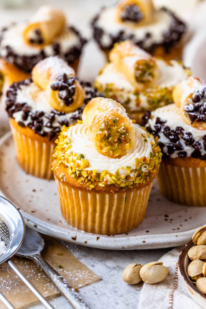 cupcakes topped with frosting and coated with topped pistachios, topped with a mini cannoli.