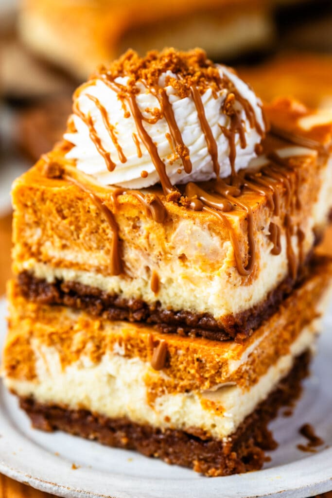 Two pumpkin cheesecake bars stacked on top of each other, with whipped cream and biscoff drizzle on top.