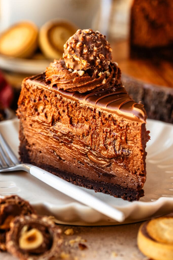 slice of Nutella cheesecake on a plate, with chocolate whipped cream and a Ferrero Rocher on top, with a fork on the side.