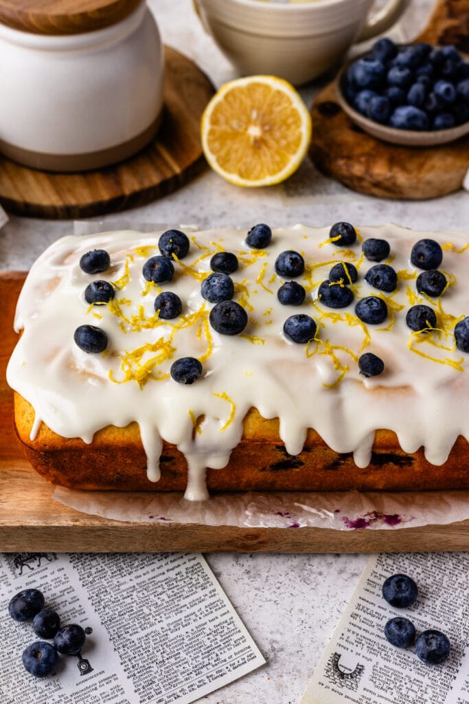 lemon blueberry load with glaze on top, and blueberries, and lemon zest, on a wooden board.